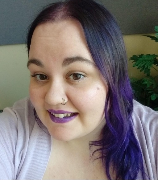 Pale feminine person with long, wavy purple hair, wearing purple lipstick at an office desk with a fake plant in the background. 
