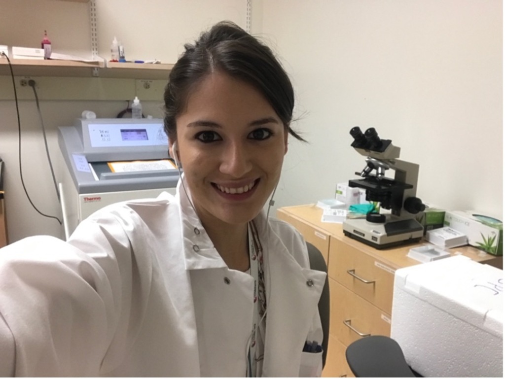 A selfie of Jennifer in the lab. She has dark hair and brown eyes. There is a microscope in the background.