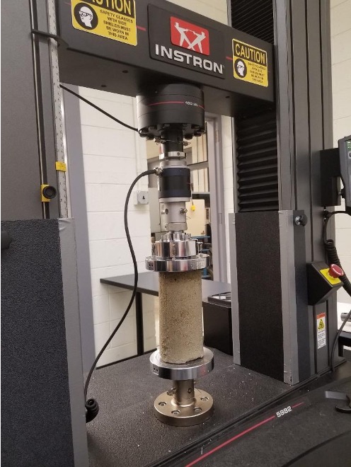 An image of a cylinder piece of cement. The cement is between two metal pieces attached to an electrical source. The top of the machine pushed down to create force, which is measured to determine the compressive strength. 